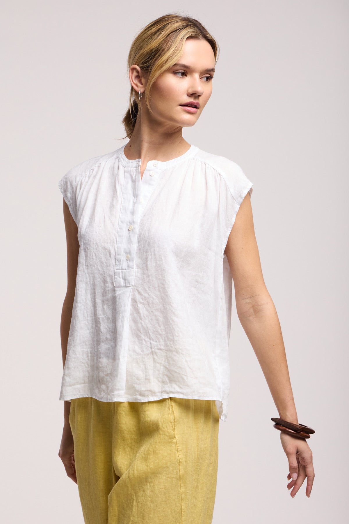 A woman wears the BEC LINEN TOP by Velvet by Graham & Spencer, a sleeveless buttoned linen piece, paired with loose yellow pants as she stands against a plain background and gazes to the side.
