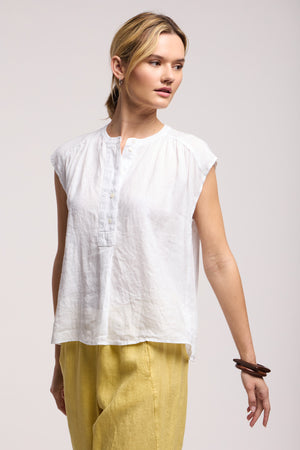 A woman wears the BEC LINEN TOP by Velvet by Graham & Spencer, a sleeveless buttoned linen piece, paired with loose yellow pants as she stands against a plain background and gazes to the side.