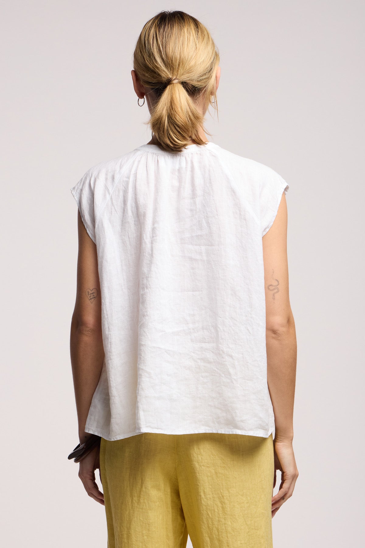 A person with blonde hair in a low ponytail is shown from the back, wearing the BEC LINEN TOP by Velvet by Graham & Spencer and yellow pants.