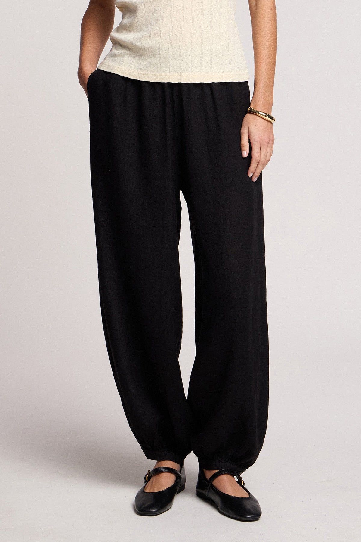 A person wears Velvet by Graham & Spencer’s CAMBER LINEN PANT—loose-fitting black linen pants with an elastic waist and cuffs—paired with a sleeveless light top, black flats, and a gold bracelet against a plain background.