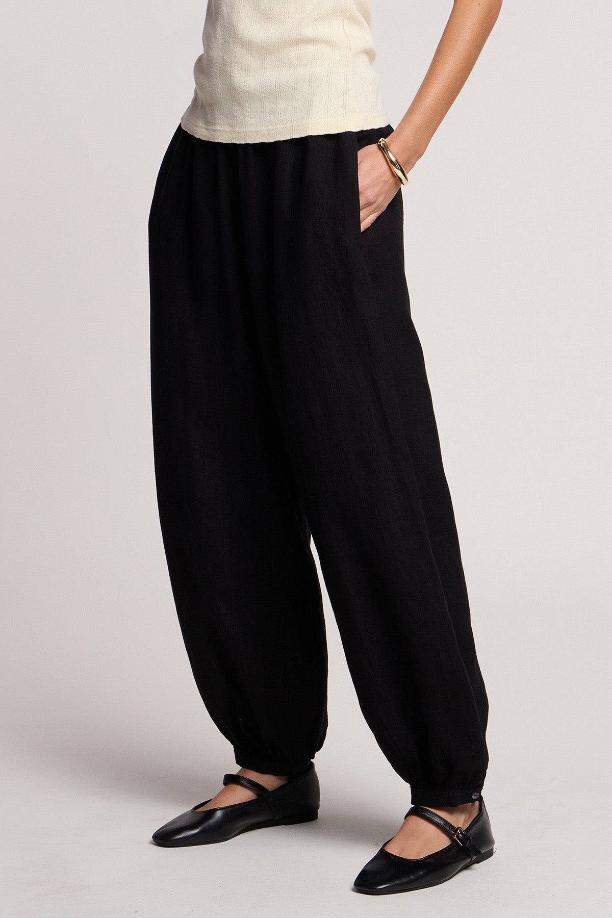 Person wearing Velvet by Graham & Spencer's CAMBER LINEN PANT in black with an elastic waist, paired with a cream short-sleeve top, black flat shoes, and a bracelet, standing hands in pockets.