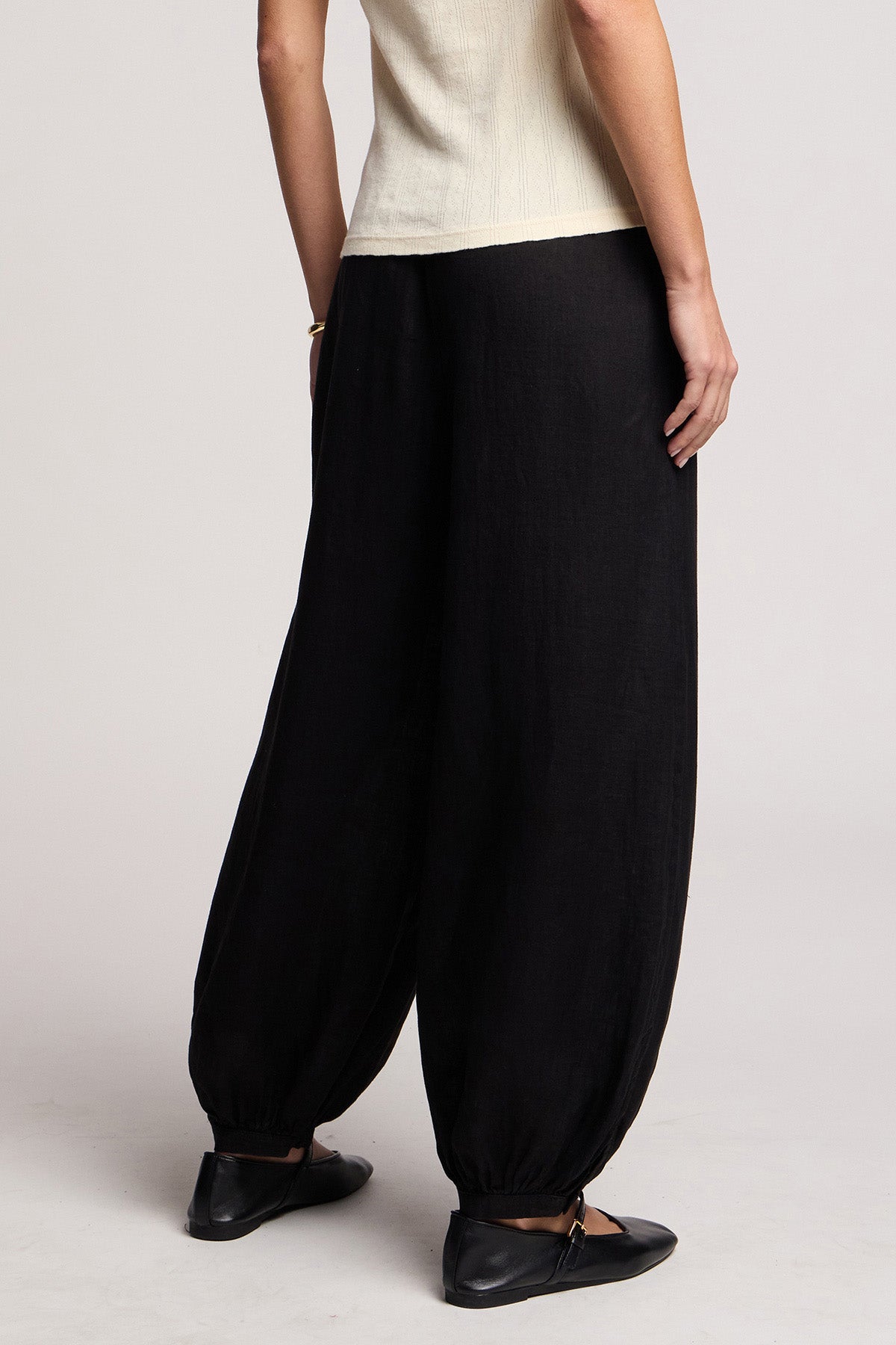 A person wears the CAMBER LINEN PANT by Velvet by Graham & Spencer—loose-fitting black linen balloon pants with an elastic waist—paired with black flats and a short-sleeved cream top, standing against a plain background.-41371564867777