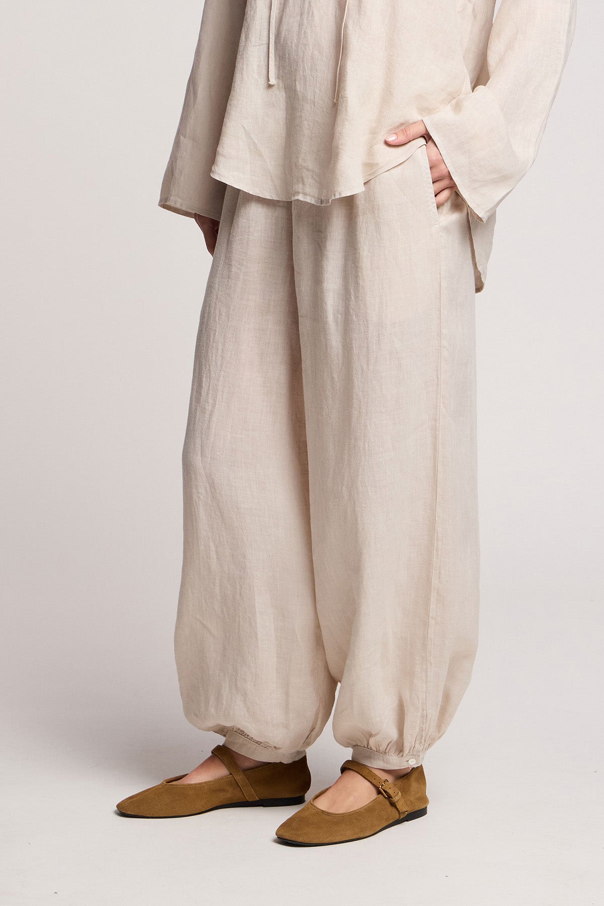 A person wears the CAMBER LINEN PANT by Velvet by Graham & Spencer—loose, light beige linen balloon pants with an elastic waist—paired with a matching woven top and brown flats, standing with one hand in a pocket.