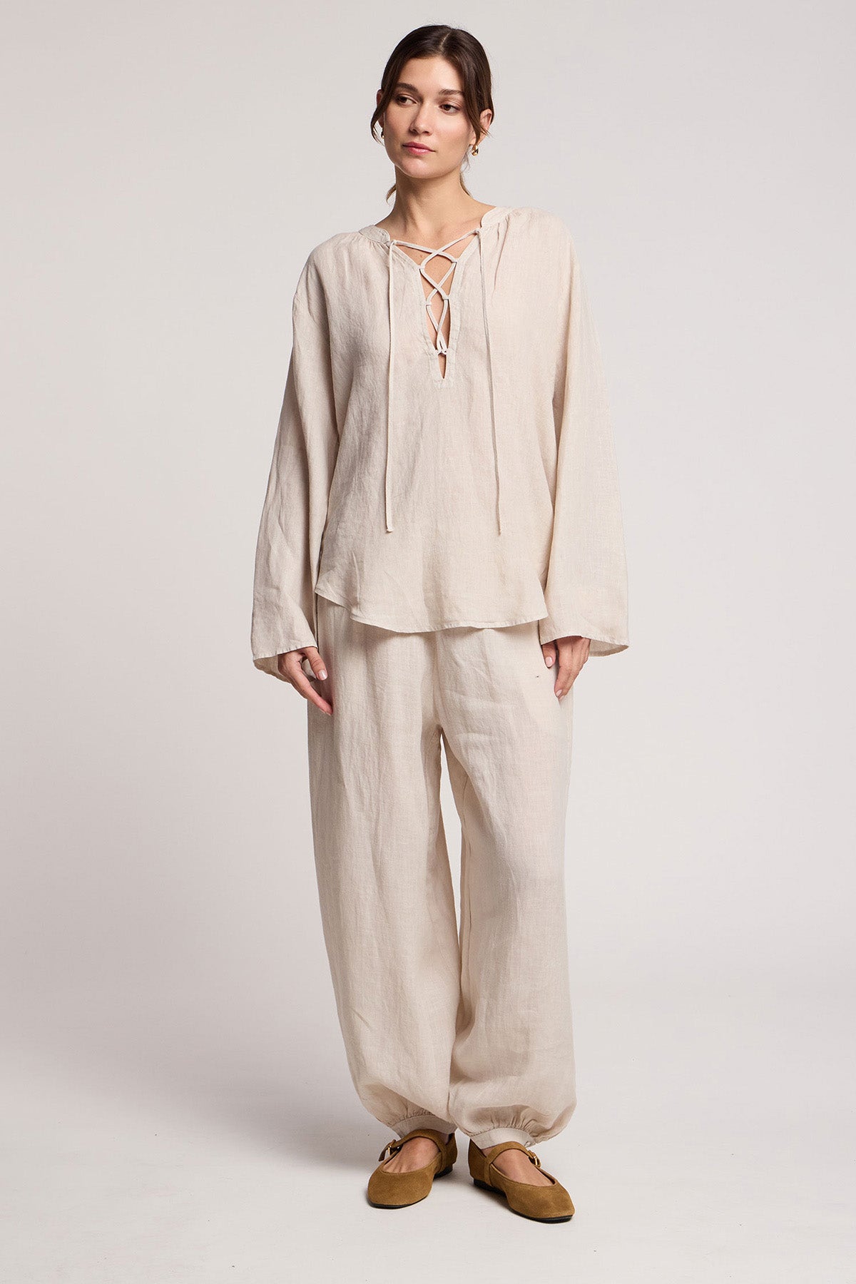 A person wearing Velvet by Graham & Spencer’s CAMBER LINEN PANT—light beige, balloon silhouette with an elastic waist—paired with a loose woven linen top featuring a lace-up neckline, against a plain light background.