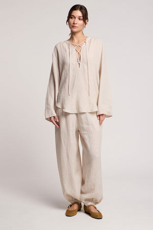 A person wearing Velvet by Graham & Spencer’s CAMBER LINEN PANT—light beige, balloon silhouette with an elastic waist—paired with a loose woven linen top featuring a lace-up neckline, against a plain light background.