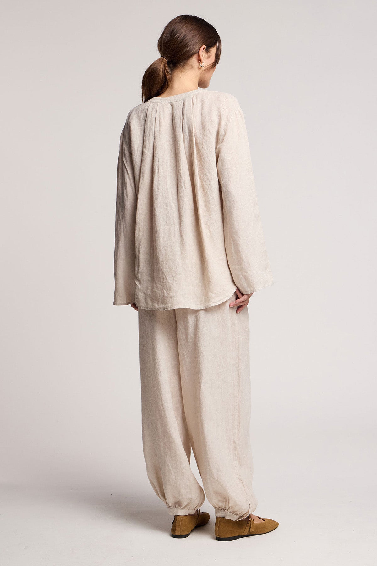 A person stands facing away, wearing a loose beige top and Velvet by Graham & Spencer CAMBER LINEN PANT with an elastic waist, paired with brown flats against a plain background.