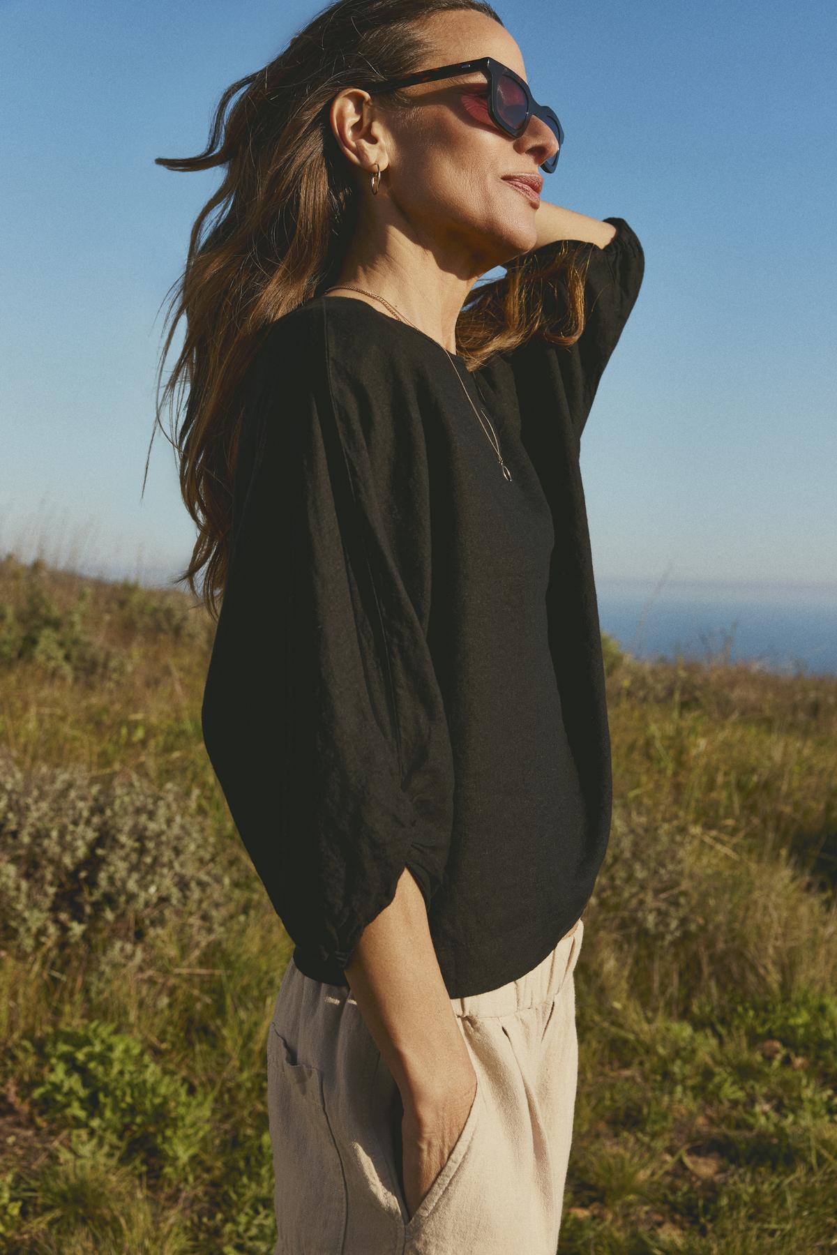 Wearing sunglasses and a Velvet by Graham & Spencer CASS LINEN TOP featuring a twist knot detail, a woman stands in a sunny field.-36443459289281