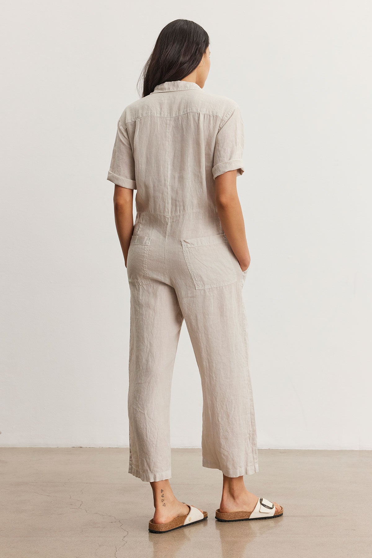   Woman standing facing away, wearing a beige short-sleeved Velvet by Graham & Spencer DELILAH LINEN JUMPSUIT and white sandals, in a minimalist room. 