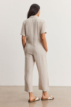 Woman standing facing away, wearing a beige short-sleeved Velvet by Graham & Spencer DELILAH LINEN JUMPSUIT and white sandals, in a minimalist room.