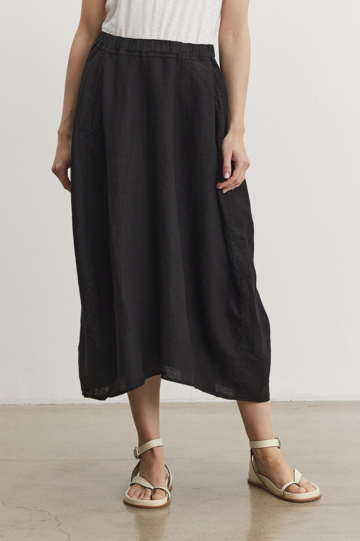   Person wearing a black FAE LINEN SKIRT by Velvet by Graham & Spencer with an elastic waistband, a white top, and beige sandals, standing on a light brown floor against a plain white background. 