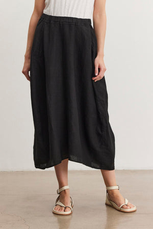 Person wearing a black FAE LINEN SKIRT by Velvet by Graham & Spencer with an elastic waistband, a white top, and beige sandals, standing on a light brown floor against a plain white background.