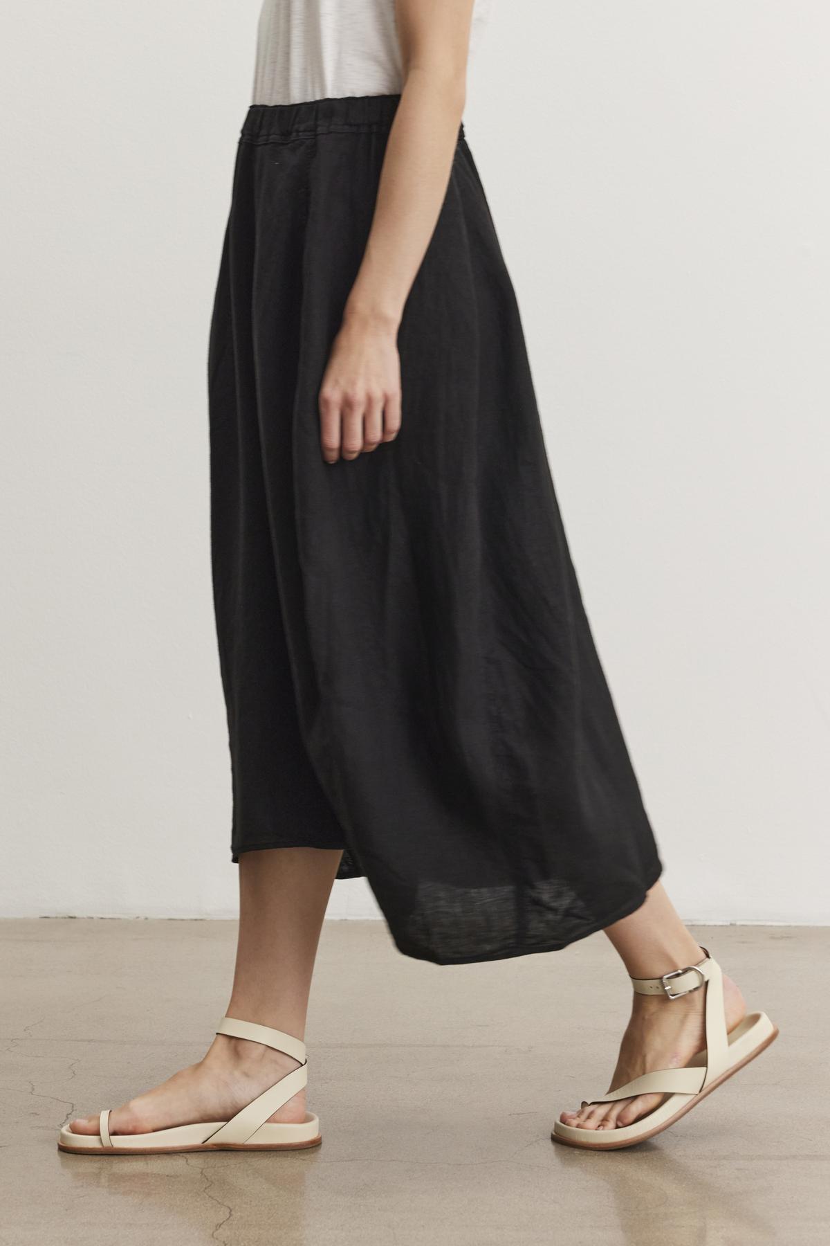   Side view of a person wearing a white top, Velvet by Graham & Spencer black FAE LINEN SKIRT with an elastic waistband, and beige sandals walking indoors on a wooden floor. 