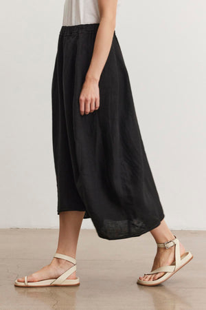 Side view of a person wearing a white top, Velvet by Graham & Spencer black FAE LINEN SKIRT with an elastic waistband, and beige sandals walking indoors on a wooden floor.