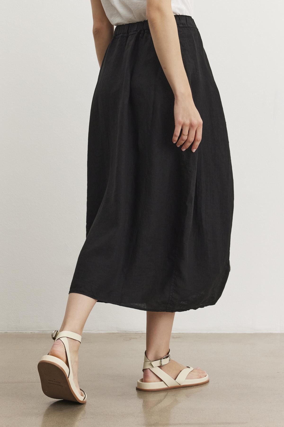 Person wearing a white top, Velvet by Graham & Spencer FAE LINEN SKIRT with an elastic waistband, and beige strappy sandals walking on a beige floor.-37104282435777