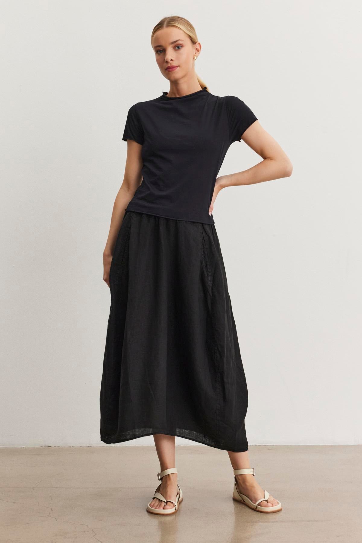 A woman stands against a plain white background wearing a black short-sleeve top, a black FAE LINEN SKIRT by Velvet by Graham & Spencer with an elastic waistband, and beige sandals. She has one hand on her hip and her feet slightly apart.-37104282337473