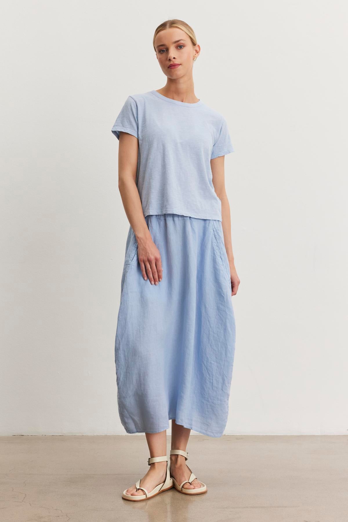   A woman stands against a plain background wearing a light blue MARIE TEE from Velvet by Graham & Spencer with a crew neckline and a matching long skirt, paired with light-colored sandals. Her outfit features a relaxed boxy fit made from breathable cotton slub. 