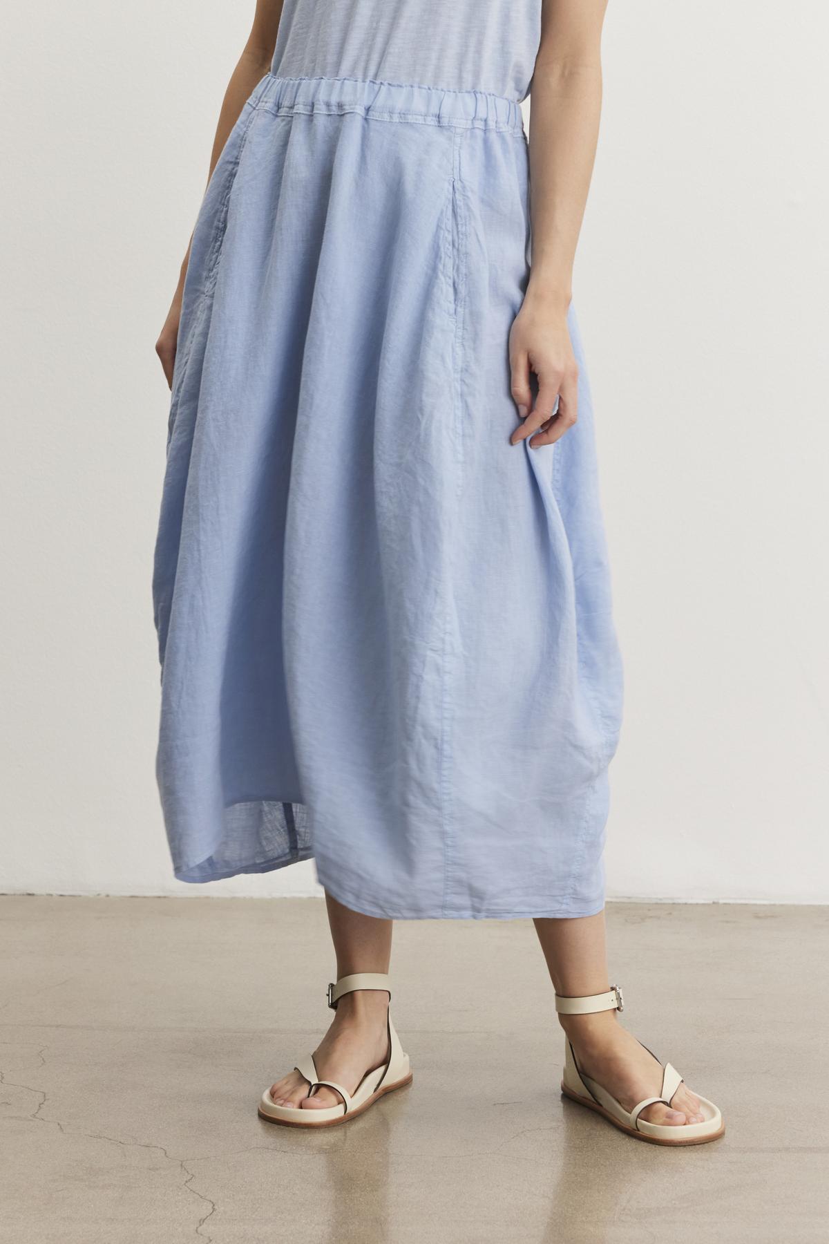   A person is standing on a polished floor, wearing a light blue, mid-length FAE LINEN SKIRT by Velvet by Graham & Spencer with an elastic waistband and beige sandals. 