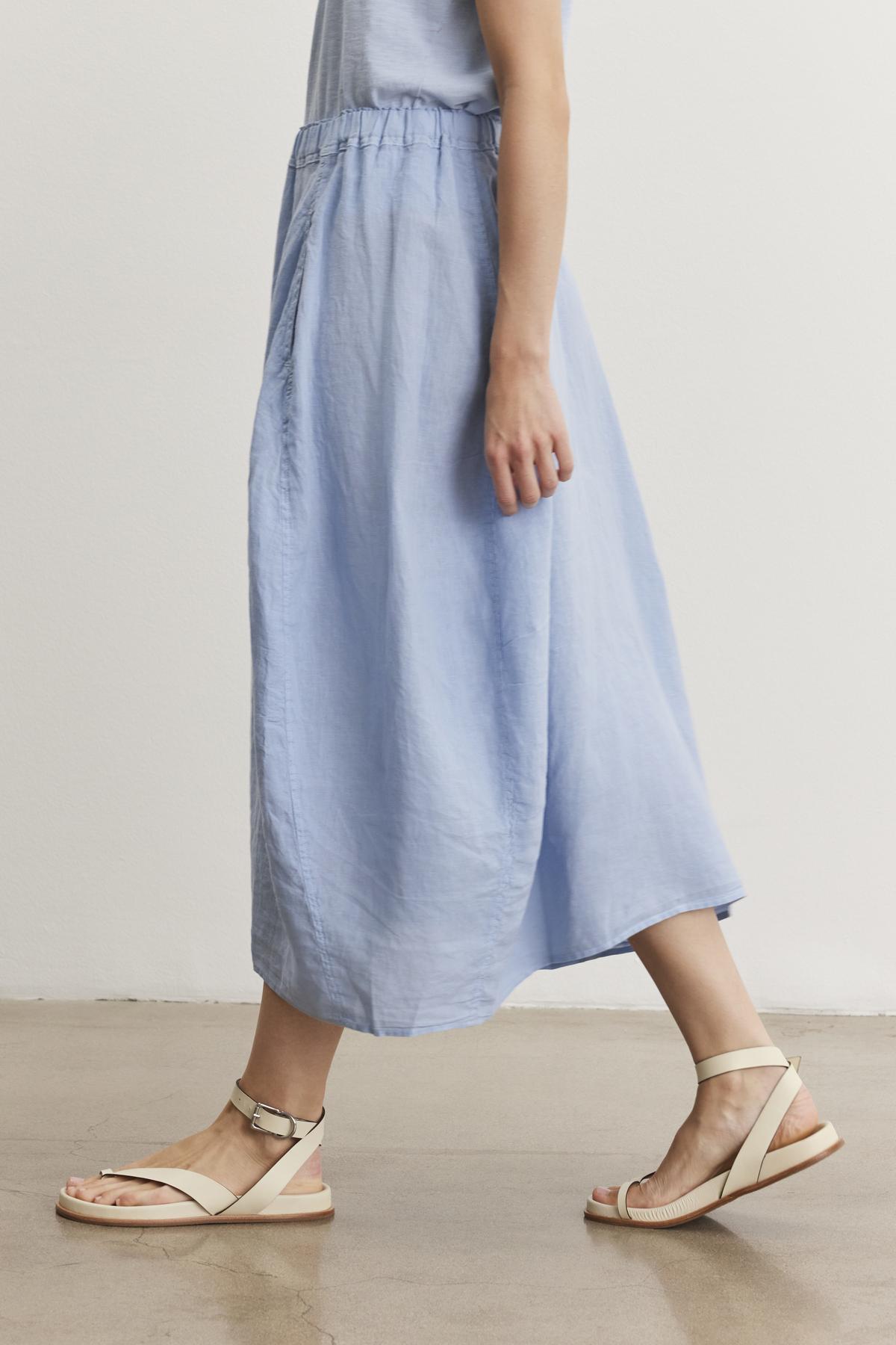   A person wearing a light blue, ankle-length FAE LINEN SKIRT by Velvet by Graham & Spencer with an elastic waistband and beige sandals is seen walking on a smooth, light brown floor. The background is a plain white wall. 