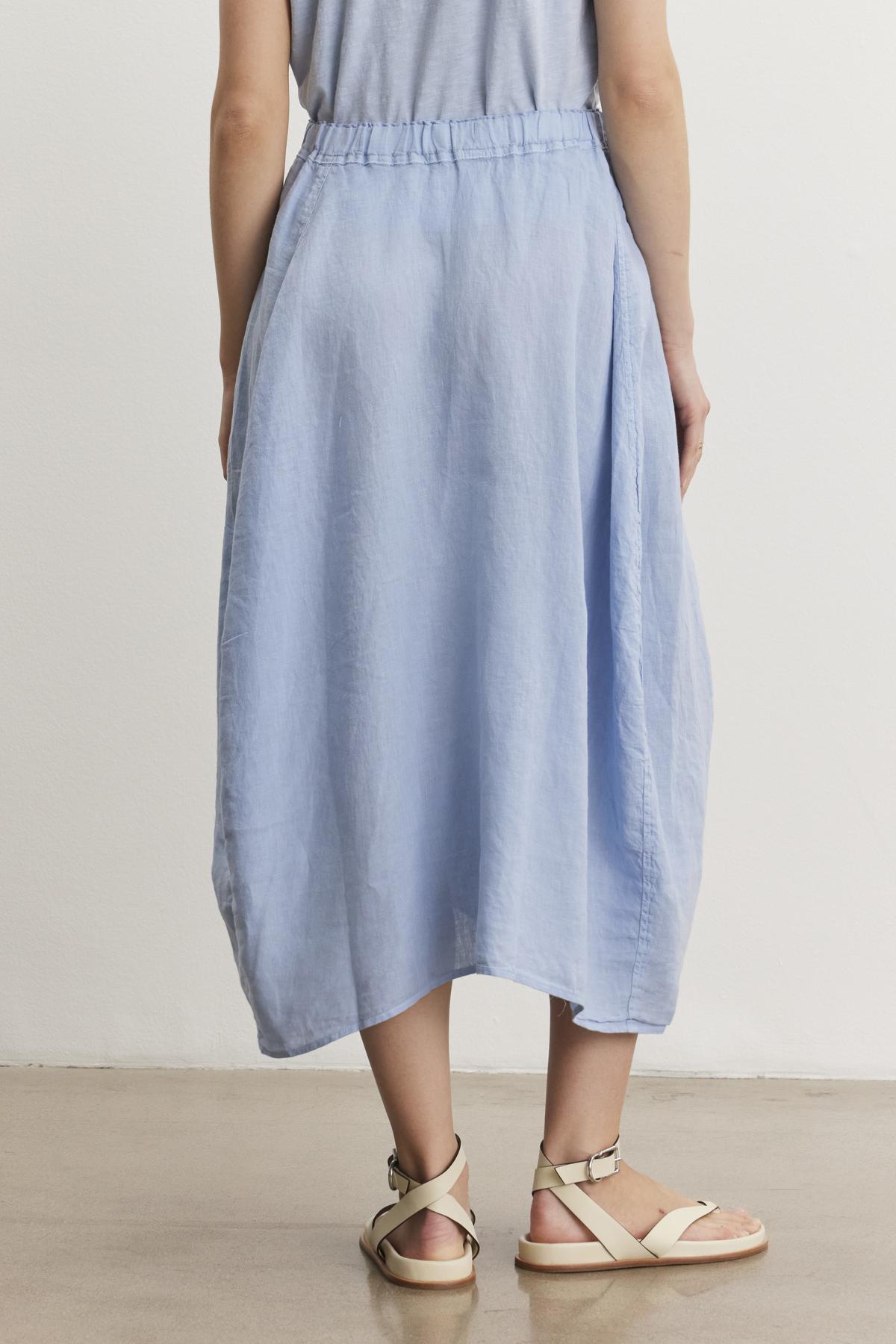   A person is standing on a smooth concrete floor, wearing a light blue, knee-length FAE LINEN SKIRT by Velvet by Graham & Spencer with an elastic waistband and light beige sandals. The top part of the outfit is partially visible. 