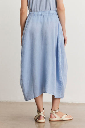 A person is standing on a smooth concrete floor, wearing a light blue, knee-length FAE LINEN SKIRT by Velvet by Graham & Spencer with an elastic waistband and light beige sandals. The top part of the outfit is partially visible.