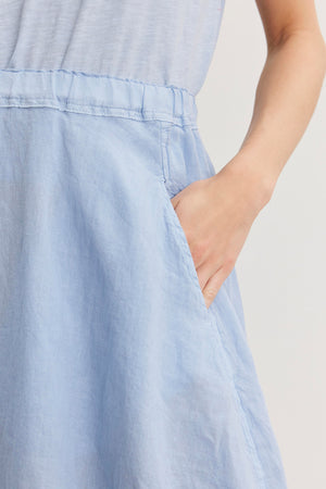 Close-up of a person’s hand in a pocket of a light blue, spring-weight linen FAE LINEN SKIRT by Velvet by Graham & Spencer. The fabric appears slightly wrinkled, and the elastic waistband gently cinches at the waist. The person's arm is partially visible.