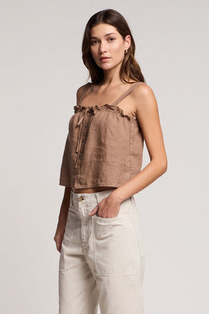 A woman stands with one hand in her pocket, wearing the IZZIE LINEN TANK TOP by Velvet by Graham & Spencer and high-waisted light-colored pants against a plain background.