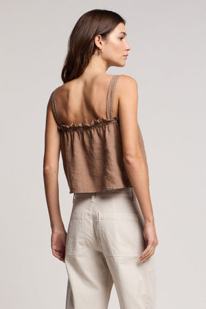 A woman stands sideways in the IZZIE LINEN TANK TOP by Velvet by Graham & Spencer—a brown woven linen cropped tank with ruffle detail—paired with high-waisted light beige pants against a plain background.