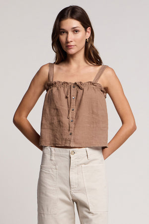 A woman wears the IZZIE LINEN TANK TOP by Velvet by Graham & Spencer—a lightweight, breathable brown woven linen cropped tank with an adjustable fit—paired with high-waisted light beige pants against a plain background.