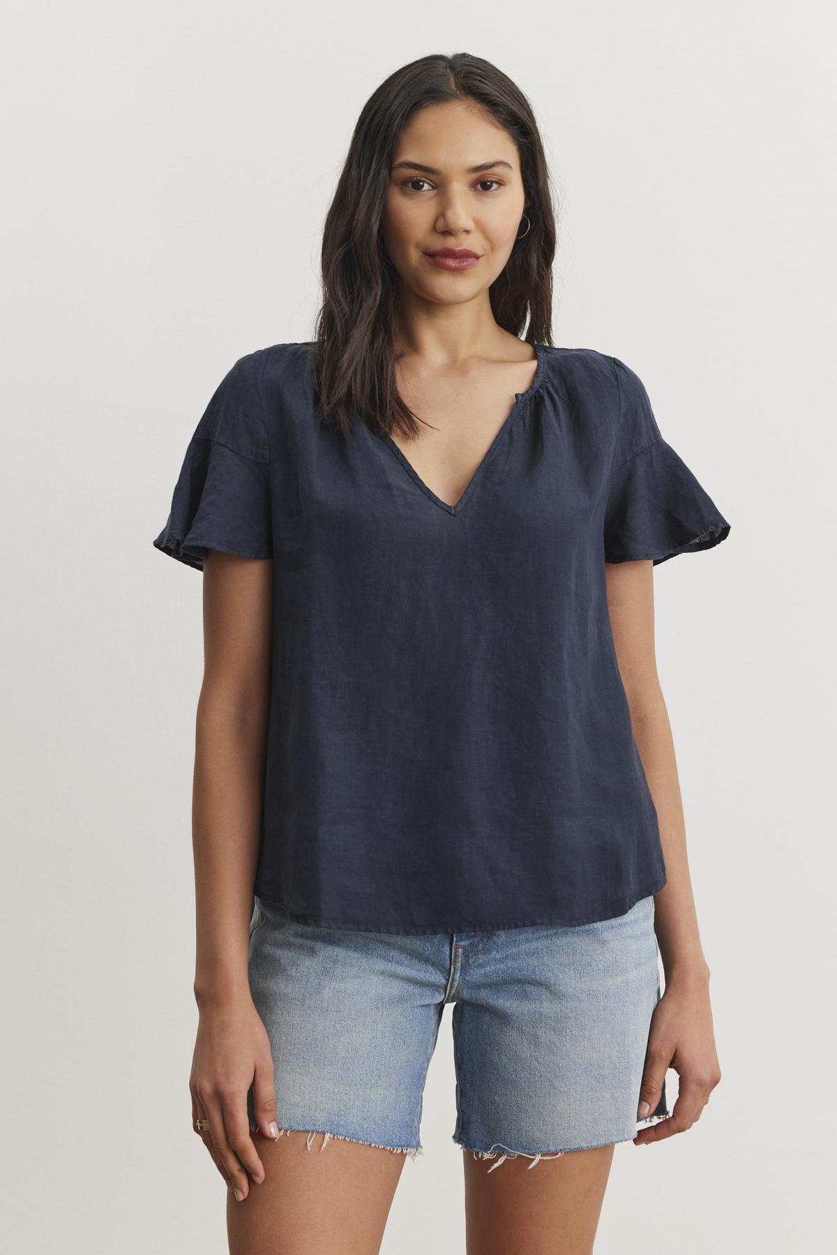   A person stands against a plain background, wearing the JENNIFER LINEN TOP in navy blue by Velvet by Graham & Spencer, paired with denim shorts. 