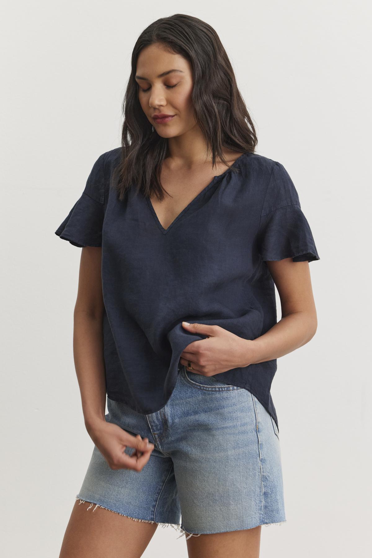   A person with long dark hair, wearing the JENNIFER LINEN TOP by Velvet by Graham & Spencer and denim shorts, stands against a plain background, looking down and slightly to the side. 