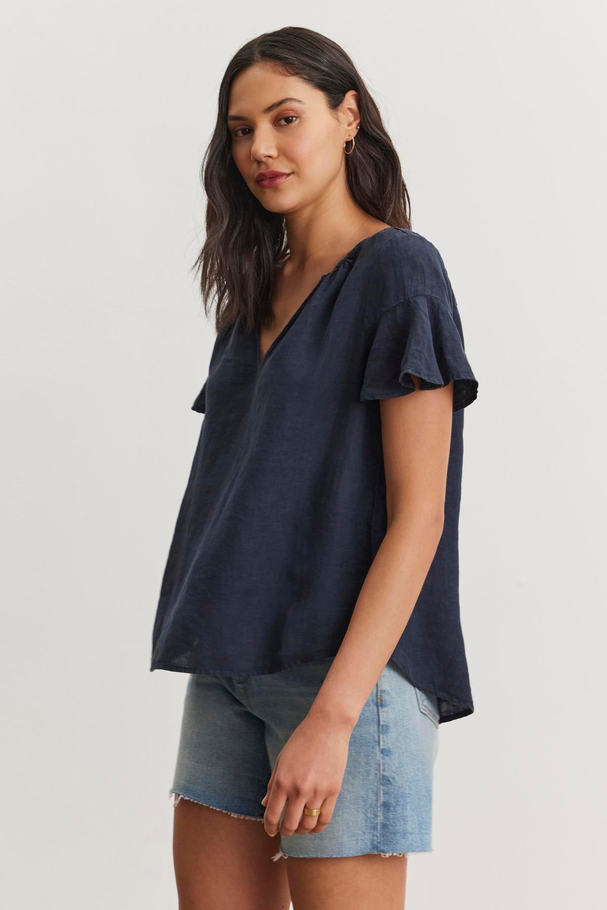   A woman with shoulder-length dark hair is wearing the JENNIFER LINEN TOP from Velvet by Graham & Spencer, which features a loose-fitting design with flutter sleeves in navy, paired with light blue denim shorts. She stands against a plain white background, highlighting the relaxed silhouette of her outfit. 