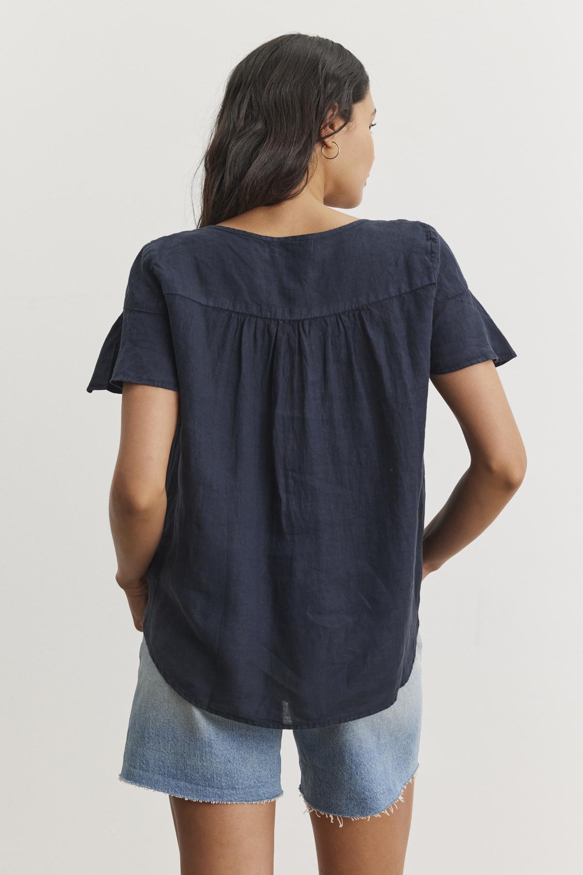   A person with long hair is standing and facing away, wearing a relaxed silhouette with the JENNIFER LINEN TOP by Velvet by Graham & Spencer in navy blue and light blue denim shorts. 