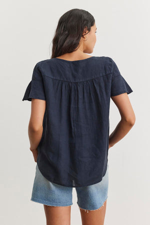 A person with long hair is standing and facing away, wearing a relaxed silhouette with the JENNIFER LINEN TOP by Velvet by Graham & Spencer in navy blue and light blue denim shorts.