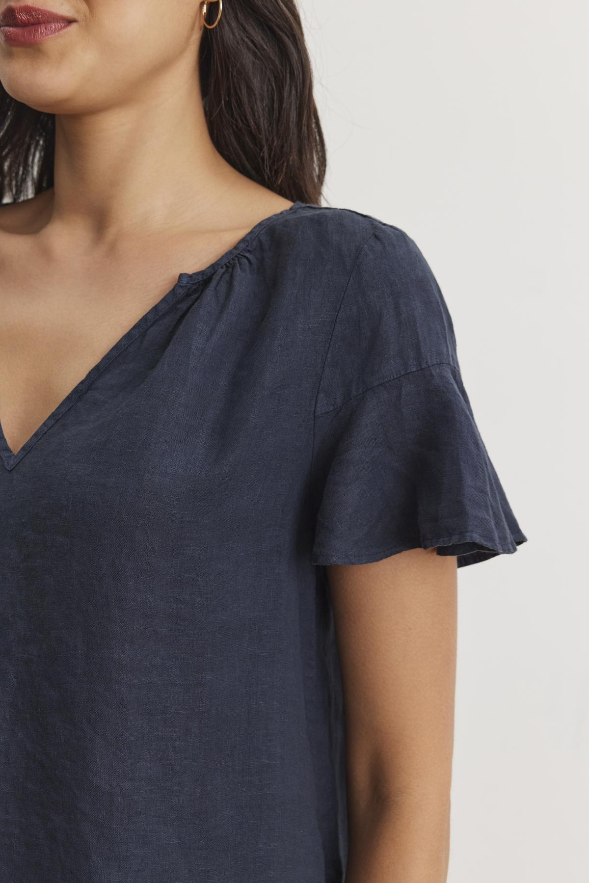   A person is wearing the JENNIFER LINEN TOP by Velvet by Graham & Spencer. This navy blue V-neck blouse features short, flutter sleeves and a relaxed silhouette that adds an air of effortless elegance against the plain white background. 