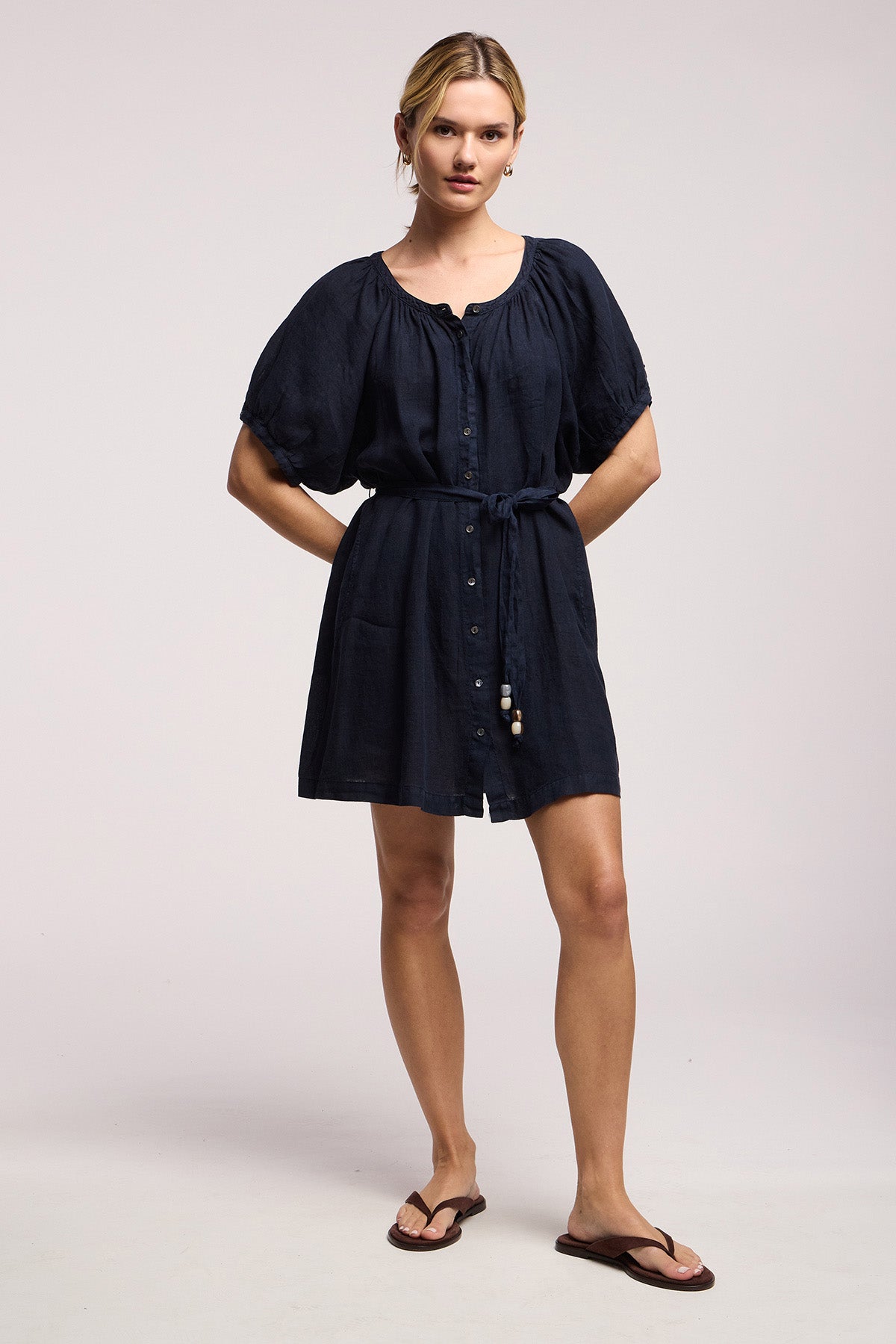 A person wears the KAYELLA LINEN DRESS by Velvet by Graham & Spencer—a short-sleeved, button-down navy tunic dress with a belted waist—paired with flip-flops, standing against a plain light background.-41527352688833