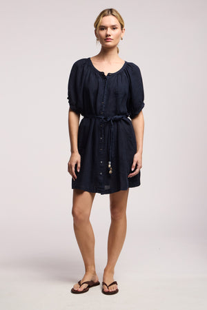 A woman stands in a studio wearing the KAYELLA LINEN DRESS by Velvet by Graham & Spencer, a navy blue, short-sleeve tunic dress with front buttons and brown flip-flops—ideal for relaxed summer style.