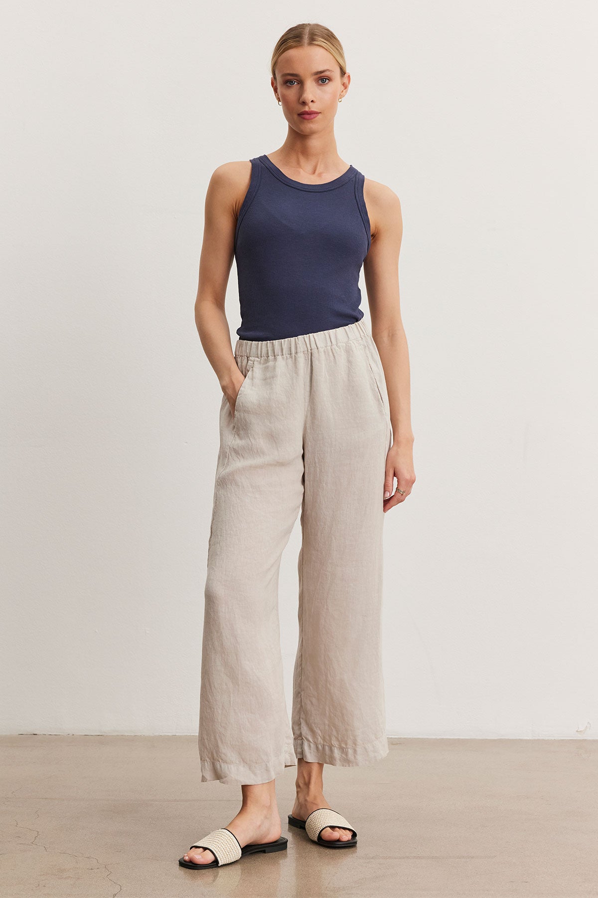 A person stands against a plain white background wearing a navy blue sleeveless top, Velvet by Graham & Spencer LOLA LINEN PANT with an elastic waist, and white slide sandals. They have their hands in their pockets.-36998848315585