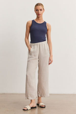 A person stands against a plain white background wearing a navy blue sleeveless top, Velvet by Graham & Spencer LOLA LINEN PANT with an elastic waist, and white slide sandals. They have their hands in their pockets.