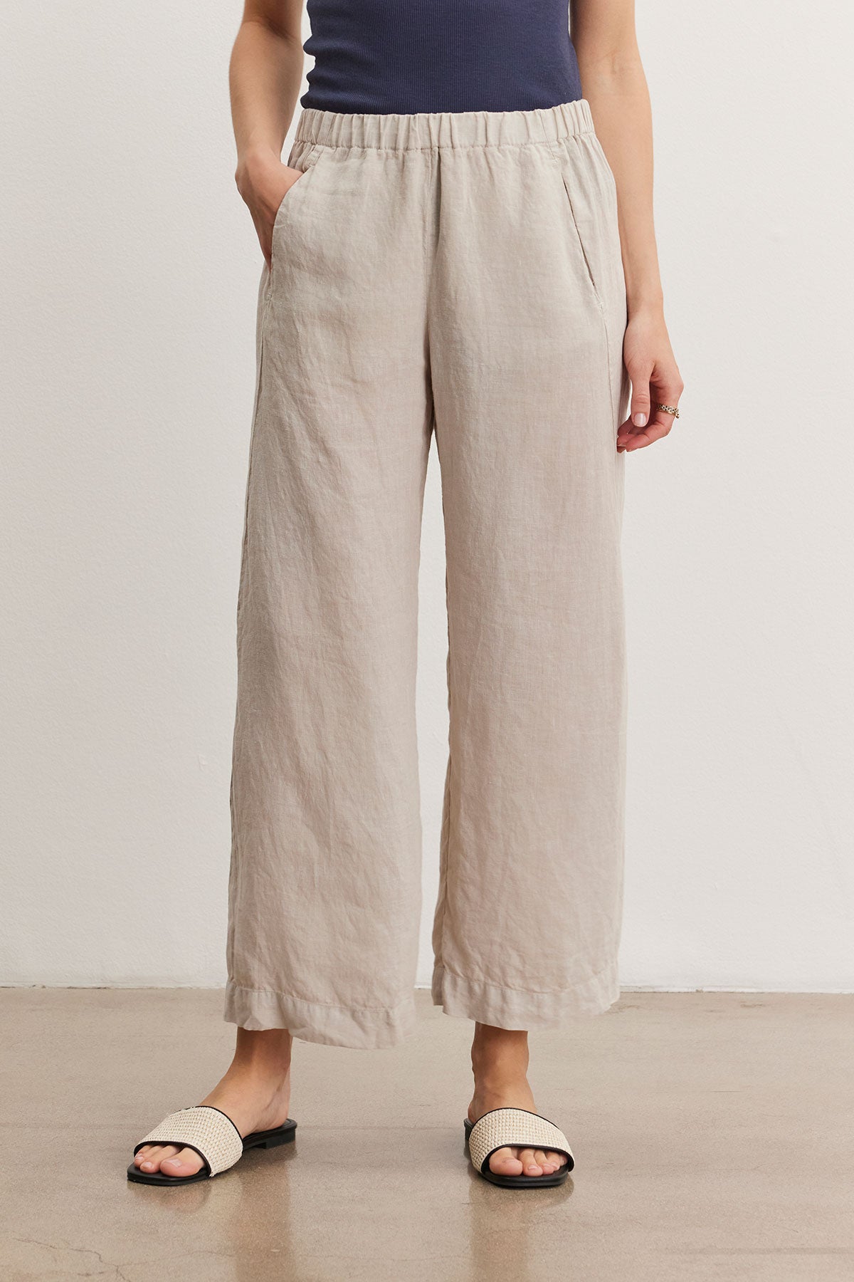   A person wearing light beige LOLA LINEN PANT by Velvet by Graham & Spencer, relaxed leg pants with hands in pockets, a dark blue top, and light-colored slip-on sandals. 