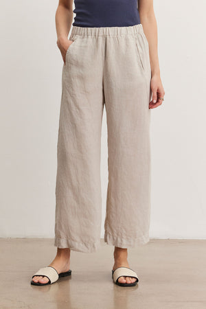A person wearing light beige LOLA LINEN PANT by Velvet by Graham & Spencer, relaxed leg pants with hands in pockets, a dark blue top, and light-colored slip-on sandals.