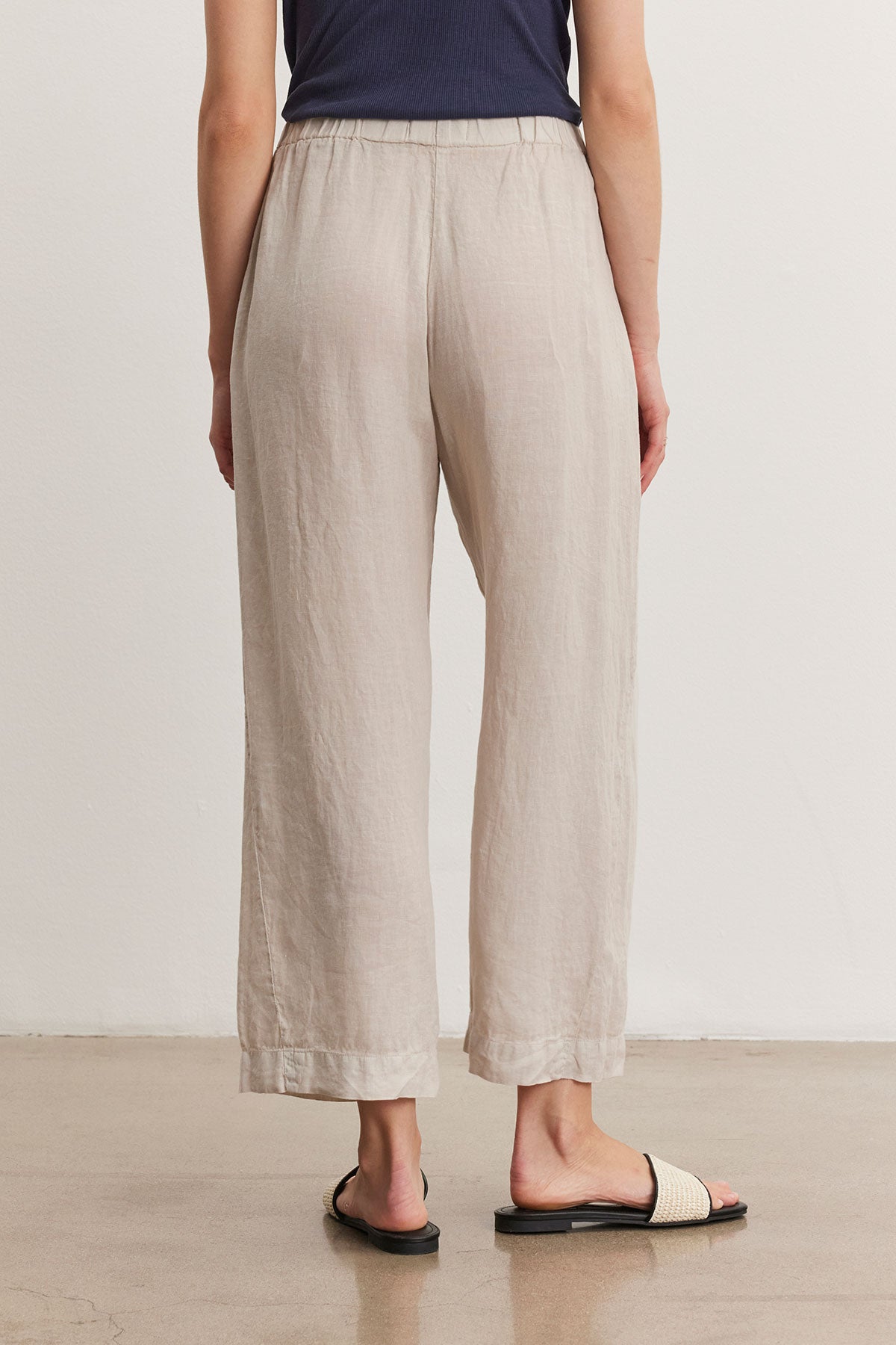   A person is standing on a polished floor, wearing beige LOLA LINEN PANT by Velvet by Graham & Spencer with an elastic waist, a navy top, and straw sandals. The photo is taken from behind. 