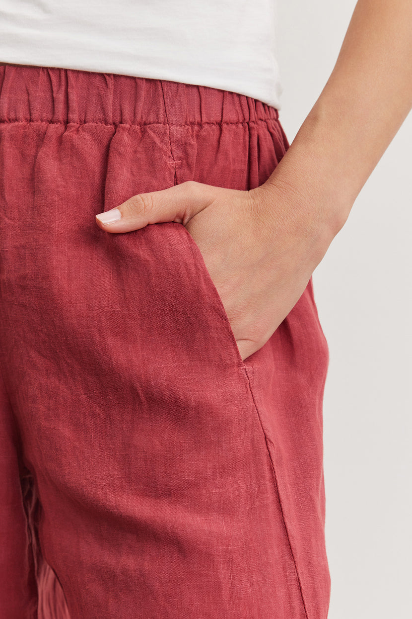 Close-up of a person's hand with a white manicure partially inserted into the pocket of an ankle crop red Velvet by Graham & Spencer Lola Linen Pant.