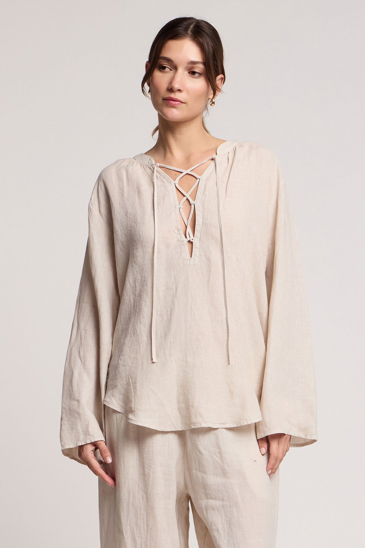 A woman wears the SALMA LINEN TOP by Velvet by Graham & Spencer—a loose-fitting, cream woven linen top with a lace-up V-neck—paired with matching wide-leg pants, standing against a plain light background.-41372115501249