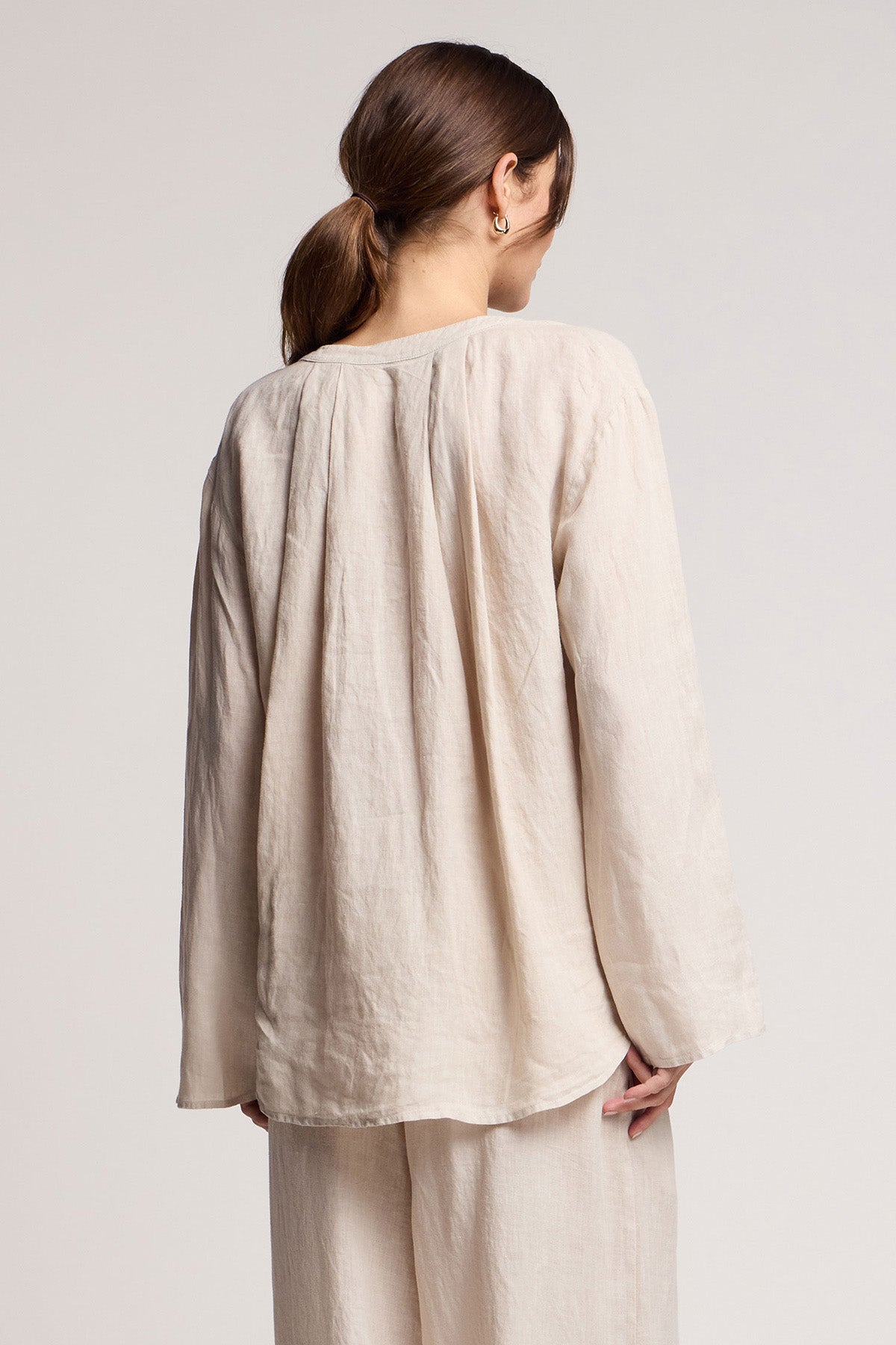 A woman with brown hair in a low ponytail faces away, wearing the Velvet by Graham & Spencer SALMA LINEN TOP and matching pants against a plain background.