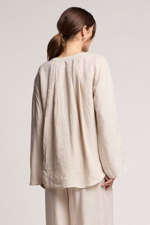 A woman with brown hair in a low ponytail faces away, wearing the Velvet by Graham & Spencer SALMA LINEN TOP and matching pants against a plain background.