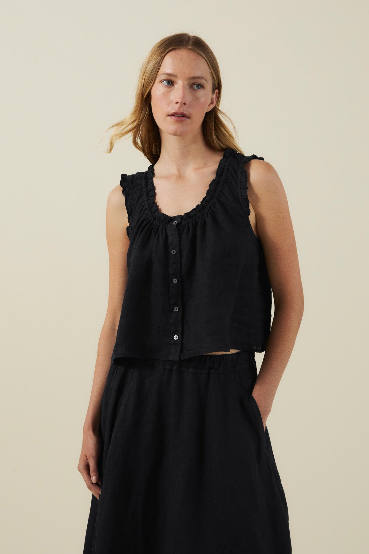 A woman wearing the TANA LINEN TOP by Velvet by Graham & Spencer—a sleeveless black button-front linen top with ruffle trim—paired with a matching black skirt, stands against a plain light background.-42118586040513