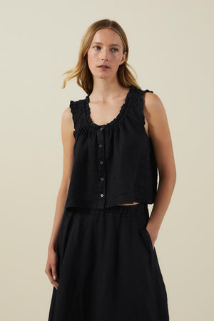 A woman wearing the TANA LINEN TOP by Velvet by Graham & Spencer—a sleeveless black button-front linen top with ruffle trim—paired with a matching black skirt, stands against a plain light background.