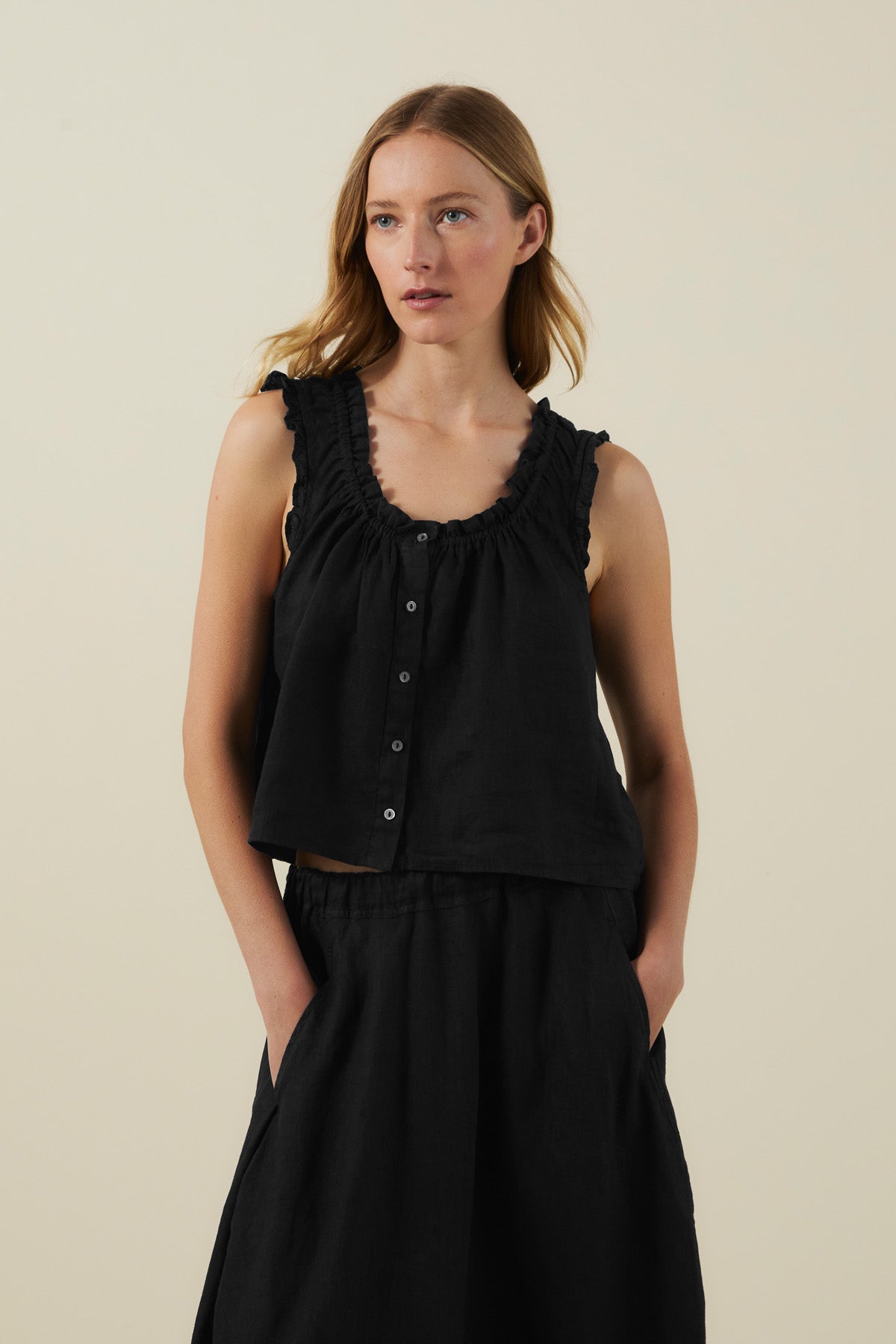 A woman wears the TANA LINEN TOP by Velvet by Graham & Spencer—a sleeveless black linen top—paired with a matching black skirt, standing with her hands in the pockets against a plain light background.-42118586007745