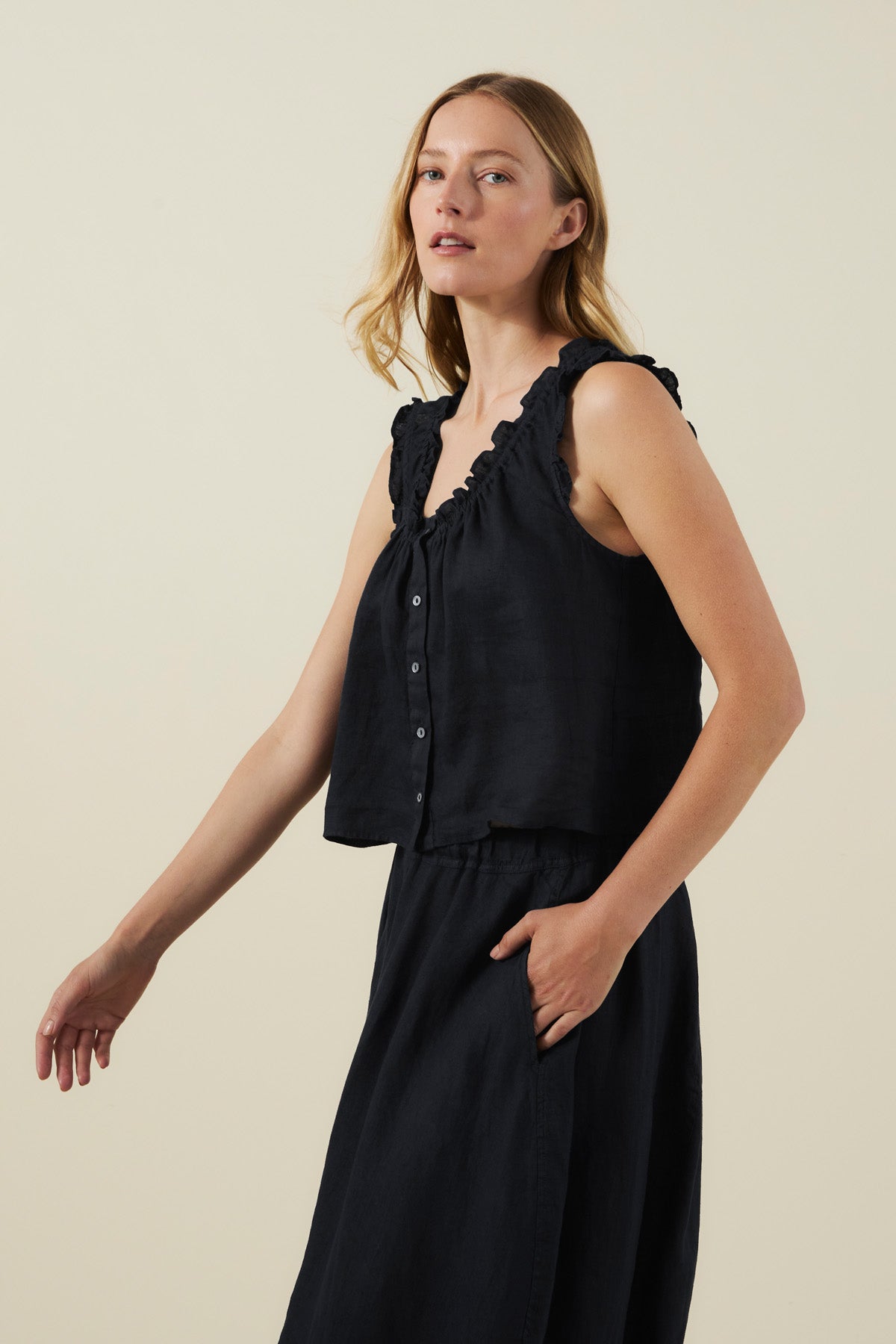 A woman with long light brown hair wears the TANA LINEN TOP by Velvet by Graham & Spencer—a sleeveless black linen top with subtle ruffle trim—paired with a matching black skirt, standing against a plain beige background.