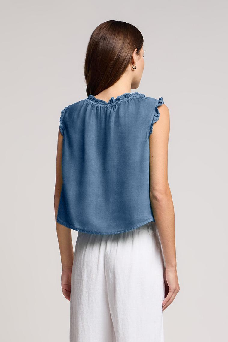 A person with long brown hair stands with their back to the camera against a plain background, wearing the TANA LINEN TOP by Velvet by Graham & Spencer—a sleeveless blue button-front linen top—paired with white pants.